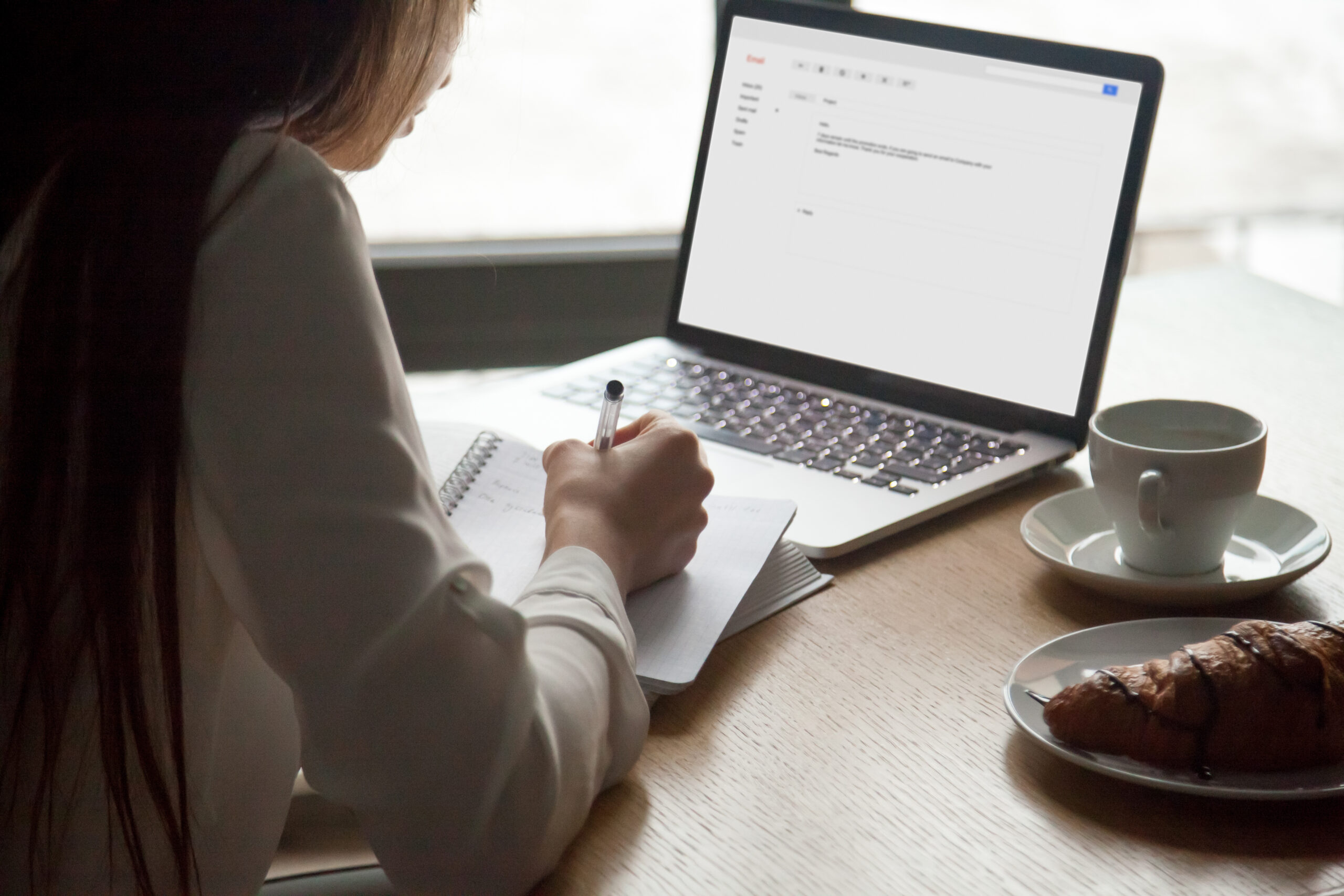 Woman making notes reading email letter on laptop in cafe